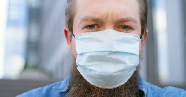 Primer plano de cara ansiosa de joven guapo hombre caucásico con barba y en máscara médica al aire libre. Retrato de un varón grave que lleva protección respiratoria en la calle. — Vídeo de stock