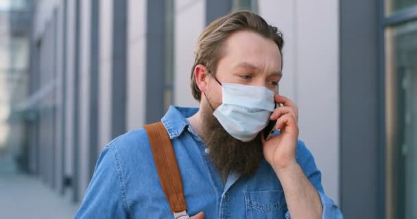 Joven caucásico con barba y máscara médica caminando por la calle y hablando por teléfono móvil. Peatón masculino en protección respiratoria paseando al aire libre y hablando por celular. Conversación . — Vídeo de stock