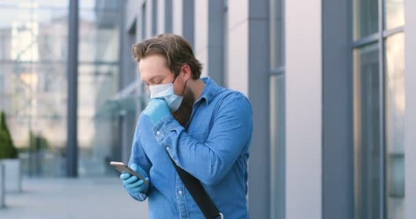 Homme piéton en masque médical marchant dans la rue, caughing et message texte sur téléphone mobile. Un homme malade en protection respiratoire se promène à l'extérieur et tape ou fait défiler sur son smartphone. — Video