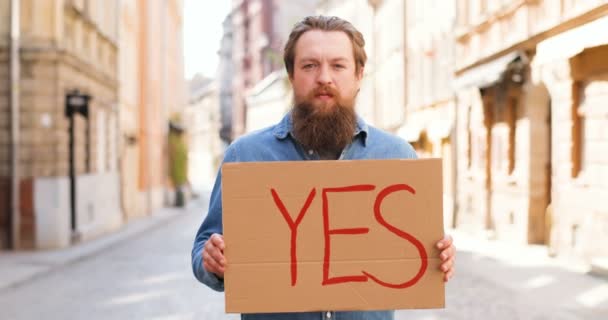 Портрет молодого кавказца, стоящего на улице в городе и показывающего плакат со словом "да". Мужчина-активист демонстрирует доску с заявлением о соглашении . — стоковое видео