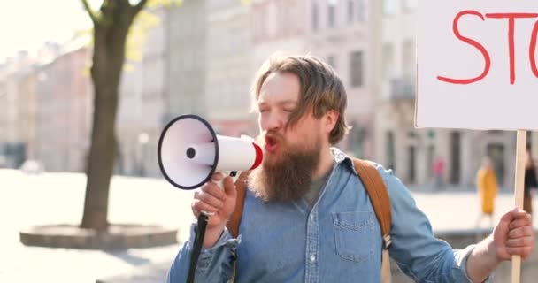 Närbild av kaukasisk manlig aktivist med skägg skrikande i megafon högtalare. Man håller affisch Stanna vid politisk eller miljömässig ensam demonstration. Single protest utomhus på torget. — Stockvideo