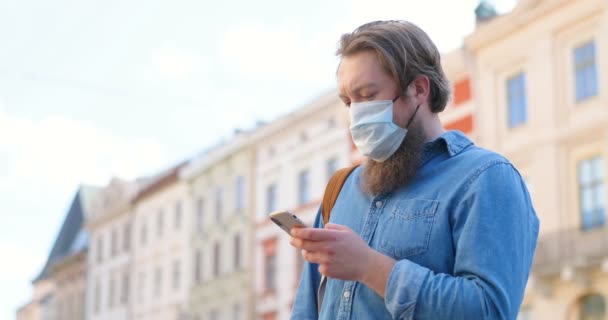 Caucasian man with beard and in medical mask standing on street and texting message on mobile phone. Male in respiratory protection is outdoor and tapping or scrolling on smartphone. Browsing online. — Stock Video