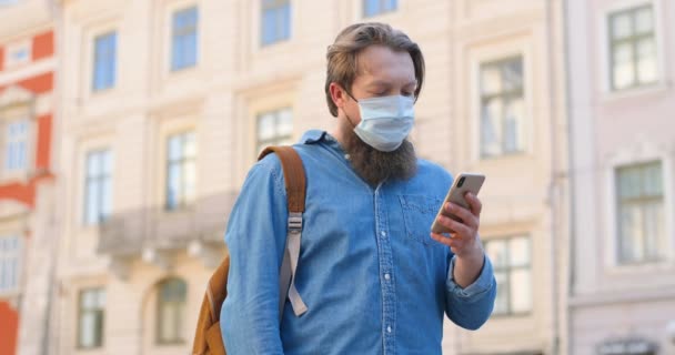 Caucasian man with beard and in medical mask standing on street and texting message on mobile phone. Male in respiratory protection is outdoors and tapping or scrolling on smartphone. Browsing online. — Stock Video