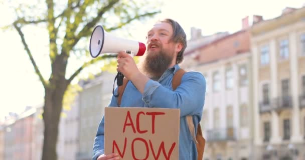 Vit manlig aktivist med skägg som talar och skriker i megafonhögtalare. Man håller affisch Act Nu på politiska eller miljömässiga ensam demonstration. Single protest utomhus på torget. — Stockvideo