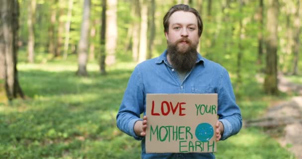 Portretfoto van blanke jonge mannelijke eco-acivist die in het groene bos staat en een poster ophangt met woorden Love our Mother Earth. Knappe man protesteert voor schone en veilige omgeving. — Stockvideo