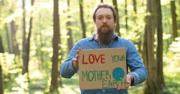 Ritratto di giovane ecologista caucasico in piedi nel verde del parco soleggiato e che tiene il tavolo con le parole Love our Mother Earth. Uomo bello con dichiarazione per la natura pulita e sicura. — Video Stock