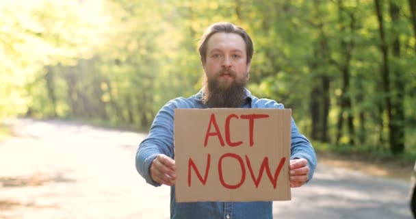 Retrato de un joven eco-acivista caucásico parado en un verde bosque soleado al lado de la carretera y sosteniendo un cartel con palabras Act Now. Hombre guapo para la naturaleza limpia y segura. Concepto de activismo ambiental . — Vídeos de Stock