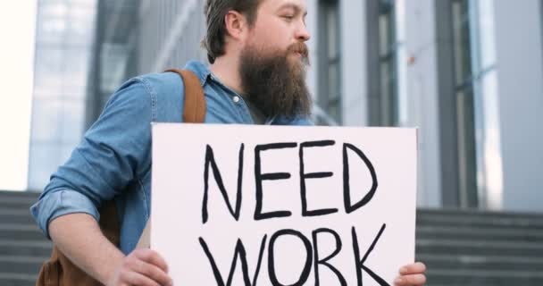 Närbild av vit ung arbetslös man med skägg står utomhus vid stora trappor och visar bord med ord Need Work. Arbetslös manlig demonstrationstavla med protest mot arbetslöshet. — Stockvideo