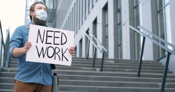 Ung vit man aktivist i medicinsk mas står på gatan i staden på stora trappor med affisch Need Work. Hårda pademiska tider. Begreppet protest mot arbetslöshet. — Stockvideo