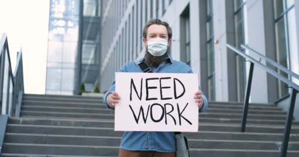 Portret van een blanke jonge werkloze man met een baard die op straat staat op een grote trap en karton toont met woorden Need Work. Mannelijke werkloze demonstratietafel met protest tegen werkloosheid. — Stockvideo