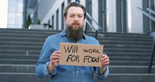 Retrato de caucasiano jovem sem trabalho homem com barba de pé ao ar livre em escadas e mostrando cartaz com palavras vai trabalhar para a comida. Homem sem emprego placa de manifestação com protesto contra o desemprego . — Vídeo de Stock