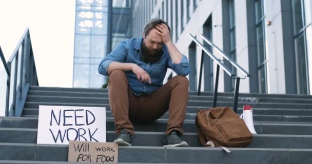 Caucásico joven triste y deprimido hombre en la desesperación sentado en las escaleras de la ciudad con carteles Necesita trabajo y Will trabajar para la comida. Hombre sin trabajo buscando ansioso y en depresión. Desempleo después del bloqueo . — Vídeos de Stock