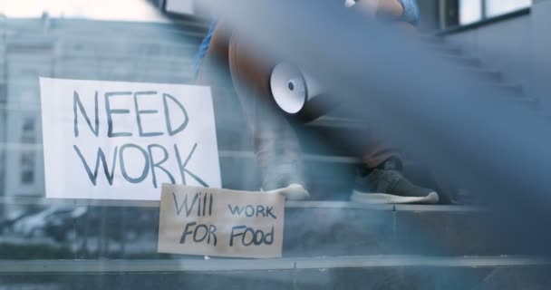 Kartónové stoly se slogany Potřebovat práci a Will pracovat pro jídlo na schodech na člověka, který sedí v zoufalství s megafonem v ruce. Mužský aktivista proti politice nezaměstnanosti. — Stock video