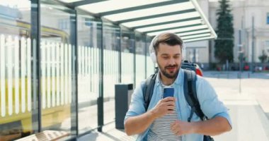 Yakışıklı, genç, beyaz, kulaklıklı gezgin otobüs durağında yürüyor ve dans ediyor. Açık havada. Çekici, mutlu erkek turist yaz günü akıllı telefondan müzik dinliyor..