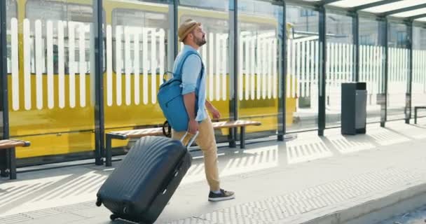 Bakre delen av kaukasisk ung stilig man i hatt och med ryggsäck promenader vid busstationen och bär resväska på hjul. Bakåt på manliga turist promenader utomhus från tågstationen eller aeroport. — Stockvideo