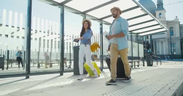 Kavkazský mladý stylový pár cestovatelů přátelé procházky na autobusové zastávce a nesoucí kufr na kolech. Pohledný muž a krásná žena, turisté procházky z vlakového nádraží nebo aeroport zdola — Stock video