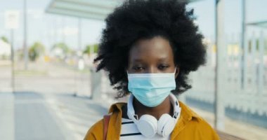Afro-Amerikan güzel bir kadının portresi. Kulaklıklı ve maskesiyle dışarıda kameraya bakıyor. Otobüs durağındaki caddede duran güzel bayana yaklaş. Kamera yüzüne yakınlaştırılıyor.