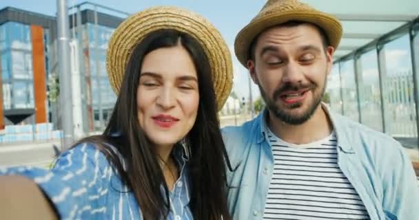 Närbild av kaukasiska unga glada par turister i hattar pratar, viftar med händerna och ler mot kameran utomhus på sommaren. POV för manliga och kvinnliga resenärer som har videochatt. Koncept för videochattning. — Stockvideo
