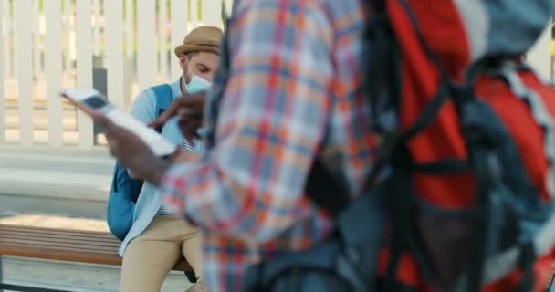Unga kaukasiska trevliga par resenärer sitter på bänken vid busshållplatsen och pratar medan de letar efter rutt på kartan. Sommarsemester. Stilig man och vacker kvinna i medalj masker med stadsplan. — Stockvideo
