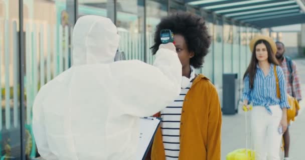Fehér védőruhát viselő személy háta, aki férfiaknál és nőknél méri a hőmérsékletet, sorban áll a szállításért. A szabadban. Férfiak és nők ellenőrzik az egészségüket az utcán. Coronavirus-járvány. Visszanézés. — Stock videók