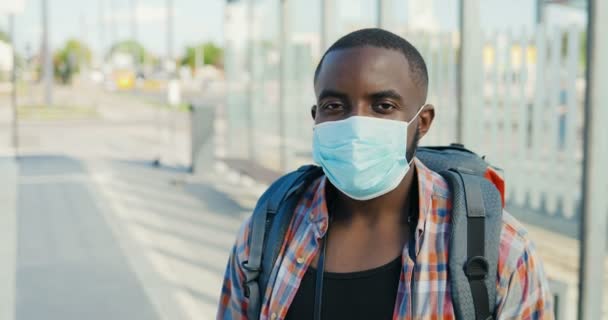 Retrato de jovem afro-americano bonito homem com mochila e em máscara médica ao ar livre olhando para a câmera. Close up de turista masculino em pé na rua na paragem de autocarro . — Vídeo de Stock