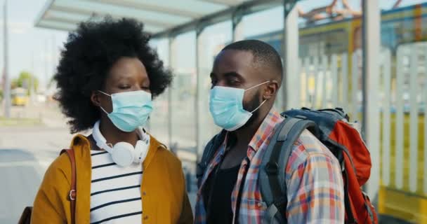 屋外のカメラを見て医療マスクで若いアフリカ系アメリカ人の観光客の肖像画。かなり幸せな女性とハンサムな男性のバス停留所で通りに立って閉じます。カメラのズーム. — ストック動画