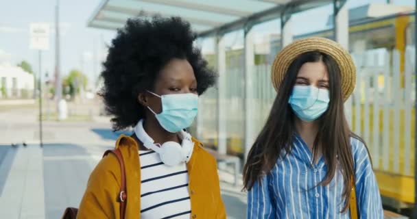 Retrato de jovem casal afro-americano de turistas do sexo feminino em máscaras médicas ao ar livre olhando para a câmera. Close up de mulheres amigas muito felizes na rua na parada de ônibus. Meninas alegres durante a pandemia . — Vídeo de Stock