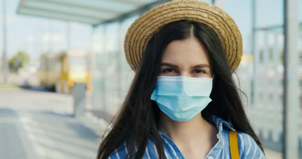 Portrait of young Caucasian beautiful woman in hat and medical mask outdoors looking at camera. Close up of pretty brunette female standing at street at bus stop. — Stock Video