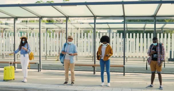 Smíšené rasy mladých mužů a žen lidé im lékařské masky stojí ve frontě na autobusové zastávce. Udržujeme si bezpečnou společenskou vzdálenost. Multiethnic stylové muži a ženy turisté venkovní čeká na dopravu. — Stock video