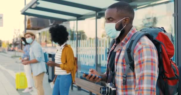 Blandade raser unga män och kvinnor människor im medicinska masker står i kö vid busshållplatsen. Hålla ett säkert socialt avstånd. African American snygg man turist utomhus väntar på transport. Turister. — Stockvideo