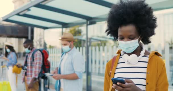 Blandade raser unga män och kvinnor människor im medicinska masker står i kö vid busshållplatsen. Hålla ett säkert socialt avstånd. African American elegant kvinna turist utomhus väntar på transport. Resenärer — Stockvideo