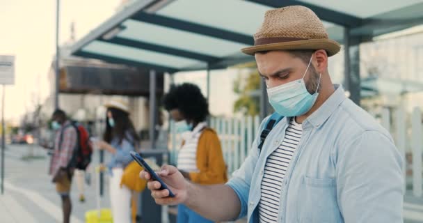 Multietniska unga män och kvinnor människor im medicinska masker står i kö vid busshållplatsen. Hålla ett säkert socialt avstånd. Vit snygg man turist utomhus väntar på transport. Turister. — Stockvideo