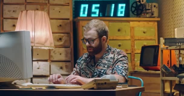 Blanke jongeman met baard, in bril en kleurrijk Hawaïaans shirt zittend in vintage kamer op PC monitor en typend op toetsenbord. Man werkt aan retro computer. Freelancer of hacker uit de jaren '80 — Stockvideo