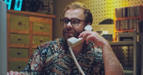 Jeune homme caucasien en lunettes et chemise colorée assis dans une pièce désordonnée des années 80. Homme parlant sur le téléphone vintage à la maison avec fil et travaillant sur ordinateur dans les années 90. Style rétro pigiste. — Video