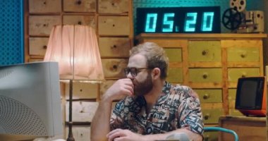 Gözlüklü ve renkli tişörtlü beyaz bir gazeteci klasik PC monitöründe çalışıyor, düşünüyor ve klavyede daktilo kullanıyor. 70 'lerin erkek yazarı retro bilgisayarın başında oturuyor. 80 'lerin biçimi.