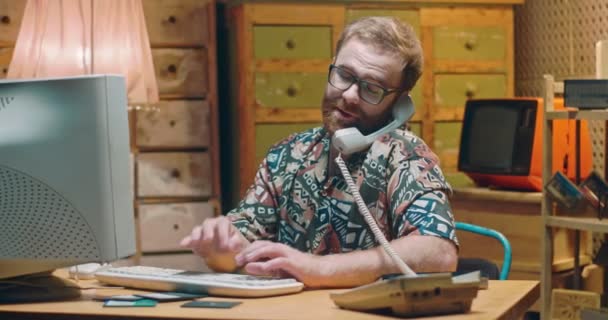 Kaukasischer junger Mann mit Brille sitzt auf einem alten PC-Monitor, tippt auf die Tastatur und telefoniert. Retro-Stil des Interieurs. Techniker oder Internetverkäufer aus den 70er und 80er Jahren am Computer. — Stockvideo