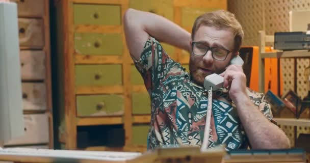 Hombre joven caucásico en gafas y camisa colorida sentado frente al monitor de PC vintage en la habitación, bostezando y llamando por teléfono. Técnico masculino trabajando en la computadora y hablando por teléfono. — Vídeo de stock