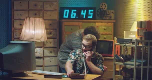 Két kaukázusi férfi programozó retro stílusban dolgozik. Egy szemüveges férfi veszi fel a régi vezetékes telefont, csörög és beszél a monitoron. 80-as vagy 90-es évekbeli férfi kockák. 70-es évek hackerei. — Stock videók