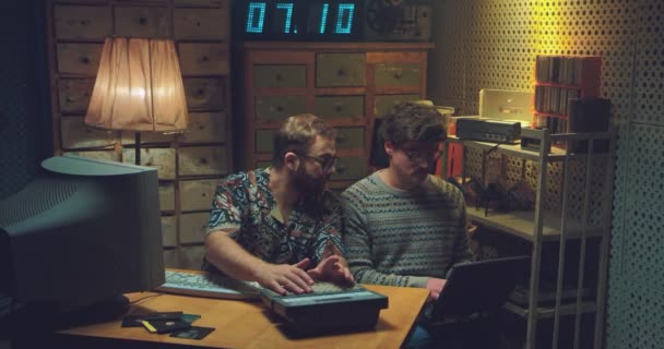 Hombres caucásicos nerds sentados en el escritorio, hablando y trabajando en la computadora. Dos programadores retro hombres escribiendo en el teclado, estudiando y contando con la vieja calculadora. Estilo vintage de los 90. Hackers de los 80. — Vídeo de stock