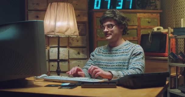 Vit manlig nörd i glasögon med mustasch sitter vid skrivbordet i retrorum och arbetar på datorn. Man programmerare skriva på tangentbordet, studera och le. Vintage stil 80-talet. Gamer från 90-talet. — Stockvideo