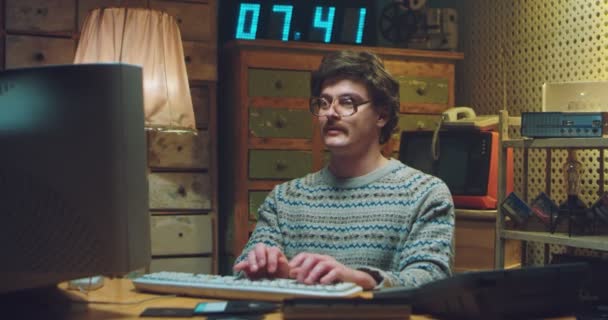 Fehér férfi stréber szemüvegben, bajusszal az íróasztalnál ül a retro szobában, és számítógépen dolgozik. Férfi programozó gépelt a billentyűzeten és beszélgetett. A 90-es évek divatos stílusa. Játékos a 80-as évekből. — Stock videók