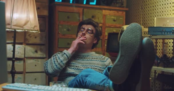Fehér férfi szemüvegben, bajusszal a 90-es években, ül a retro számítógépnél, figyel, mosolyog és cigarettázik. Lábak az asztalon. Rossz modor. Lábak az asztalon. 80-as évekbeli férfi hacker. Vintage stílus. — Stock videók