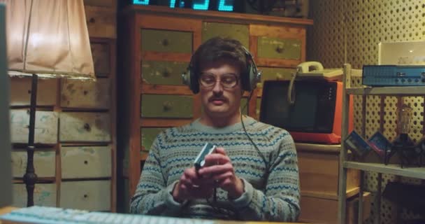 Jeune homme caucasien drôle nerd dans des lunettes et avec une moustache assis au bureau dans une salle rétro dans un casque et écoutant de la musique sur un lecteur vintage. Homme des années 80 ou 90. — Video