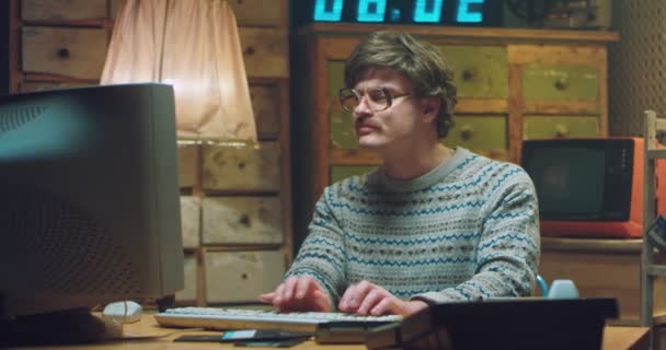 Portrait of Caucasian male goofy nerd in glasses with mustache sitting at desk in retro room and working on computer. Funny man programist looking at camera and typing on keyboard. Vintage style. — Stock Video