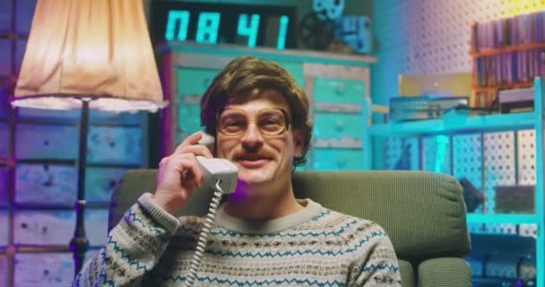 Caucásico divertido joven nerd en gafas y con bigote sentado en casa de estilo retro y hablando en el teléfono vintage cableado de los años 80. Hombre riendo y hablando por teléfono. Chico bobo de los 90. — Vídeos de Stock