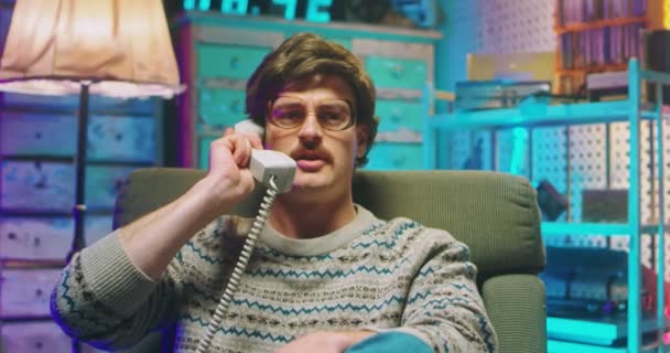 Caucasian young man nerd in glasses and with mustache sitting in retro style home and talking on wired vintage telephone of 80s. Angry stressed male speaking in phone call. Goofy guy of 90s. — Stock Video