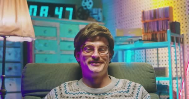 Portrait of young handsome Caucasian man nerd in glasses and with mustache sitting in armchair in retro interior room and smiling cheerfully to camera. Close up of happy male goofy from 80s. Vintage. — Stock Video