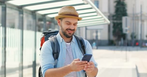 Jovem e bonito caucasiano de chapéu com mochila mensagem de texto no smartphone e sorrindo alegremente na estação ferroviária. Viajante do sexo masculino atraente tocando e rolando no telefone celular na parada de ônibus. — Vídeo de Stock