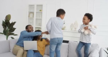 Küçük tatlı Afro-Amerikan çocuklar, kız ve erkek oturma odasında kanepede zıplayıp oynuyorlar. Ebeveynler gülümsüyor ve dizüstü bilgisayarla koltukta oturuyorlar. Çocuklar evde eğleniyor. Anne ve baba..