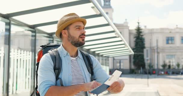 Jonge knappe blanke mannelijke toerist met hoed en rugzak staat bij de bushalte op de zomerdag met paspoort en ticket, op zoek naar weg. Goed uitziende man reiziger op het station of de luchthaven. Buiten — Stockvideo